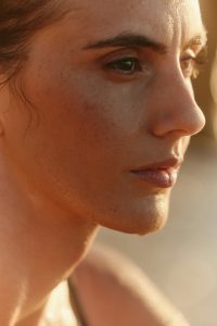 Close-up of a woman’s face with a neutral expression and visible sweat on her forehead, suggesting heat and emotional fatigue.